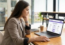 Errores comunes al usar servicios de impulso (y cómo evitarlos) Una joven profesional de marketing o emprendedora, vestida con un blazer de cuadros, sentada en un escritorio de madera en un espacio de co-working moderno y bien iluminado. Mira con concentración la pantalla de su laptop abierta, que muestra la página de "Nuestros Servicios" del sitio web Instante.net con diferentes opciones de planes. Su mano derecha está sobre el mouse, en actitud de tomar una decisión o hacer clic para seleccionar un servicio de impulso.